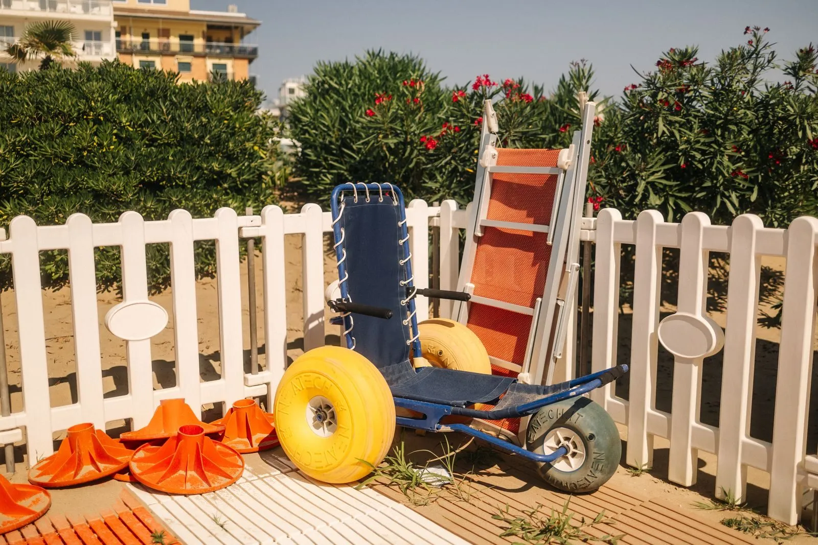Sedia da spiaggia con ruote a palloncino appoggiata alla staccionata bianca di uno stabilimento, con salvagenti e oleandri