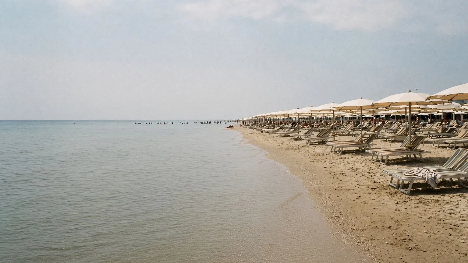 Una spiaggia attrezzata italiana vista dalla riva: diverse persone stanno in piedi nell'acqua a molti metri dalla battigia, piccole e lontane, mentre le file di ombrelloni e i lettini a doghe occupano il margine destro della sabbia