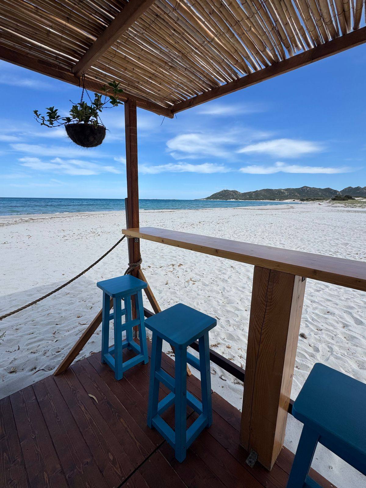 Il bancone in legno di un chiosco da spiaggia con gli sgabelli azzurri, visto da dentro verso la sabbia bianca e il mare