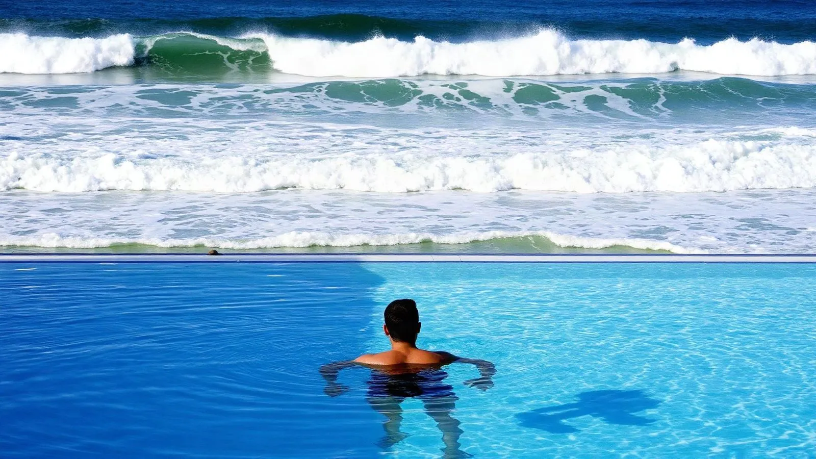 Mare mosso con le onde bianche oltre il bordo e la vasca calma dello stabilimento in primo piano, con un bagnante in acqua visto di spalle