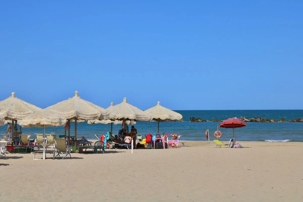 Spiaggia abruzzese con gli ombrelloni di paglia, la sabbia larga e il mare calmo dietro la scogliera frangiflutti