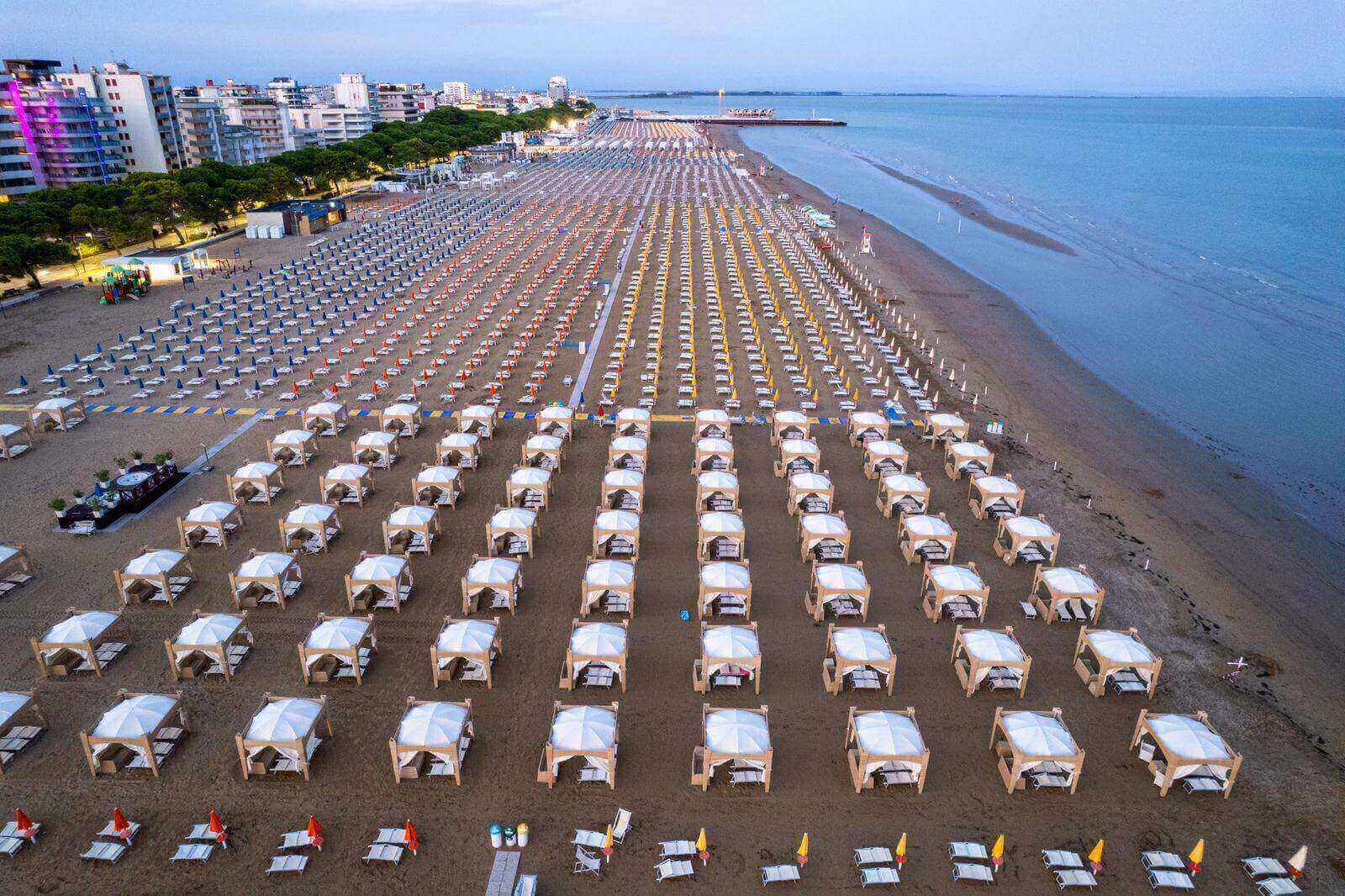 Spiaggia italiana vista dall'alto: file ordinate di gazebo bianchi e ombrelloni colorati fino alla battigia, con il lungomare e la pineta sullo sfondo