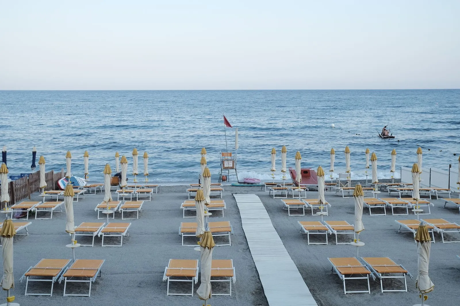 Lettini e ombrelloni chiusi allineati sulla sabbia di uno stabilimento italiano poco prima dell'apertura, con la passatoia bianca al centro e il pattino di salvataggio in riva