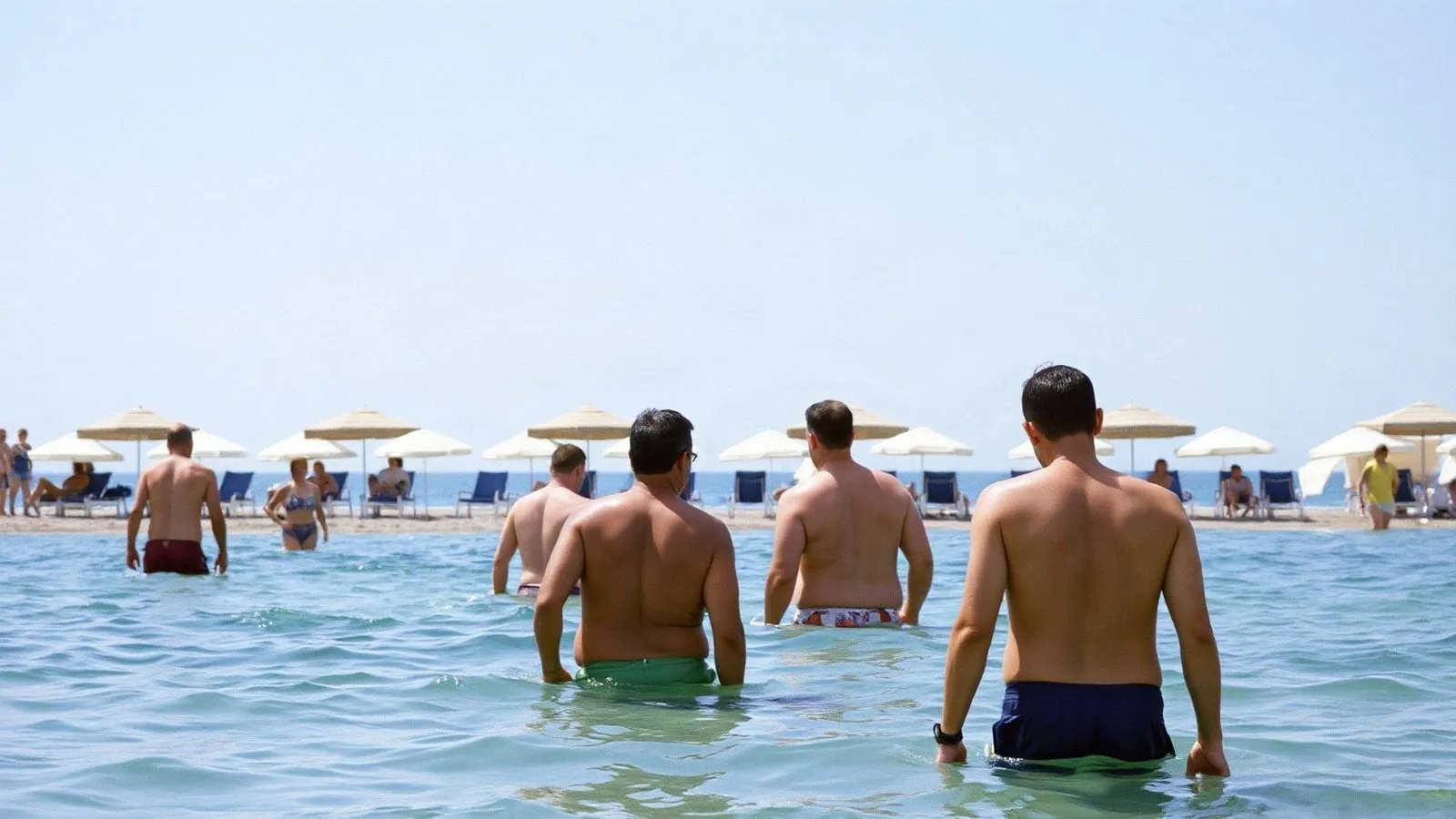 Bagnanti in piedi nell'acqua bassa a decine di metri dalla riva, visti di spalle, con gli ombrelloni dello stabilimento sullo sfondo