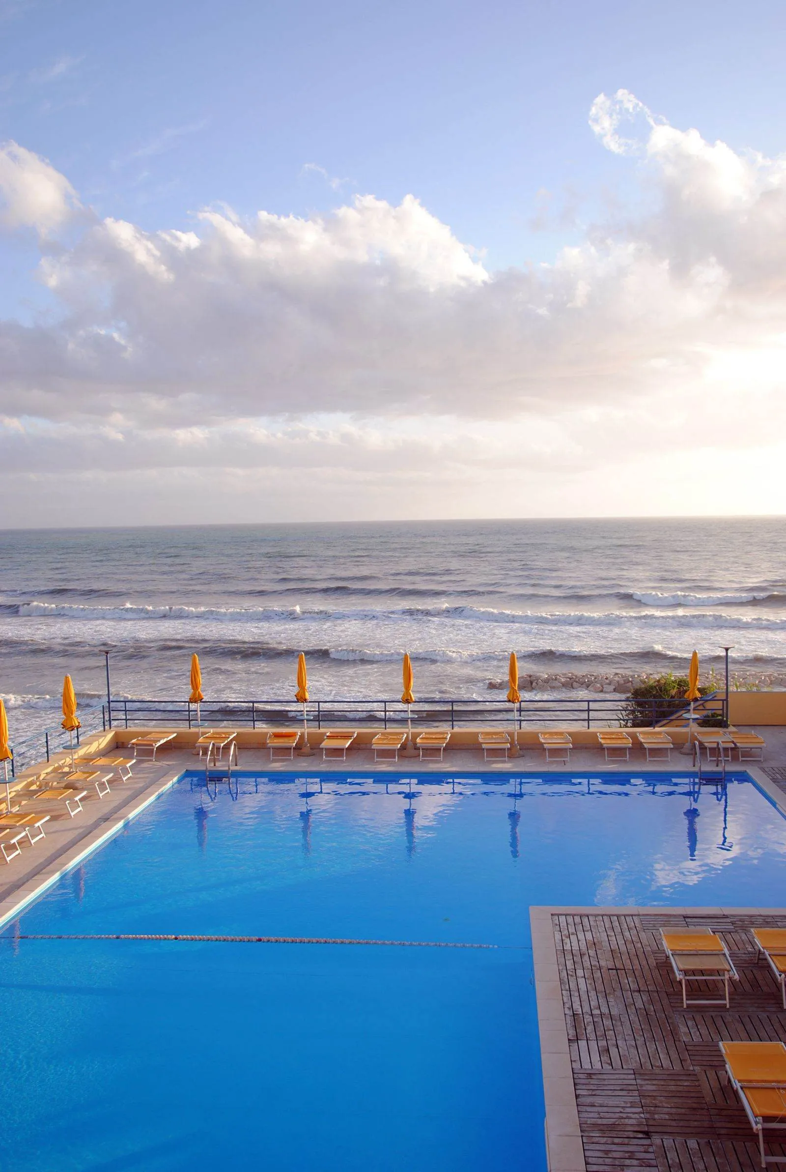 La piscina di uno stabilimento sul litorale laziale con i lettini arancioni intorno e il mare mosso oltre la ringhiera