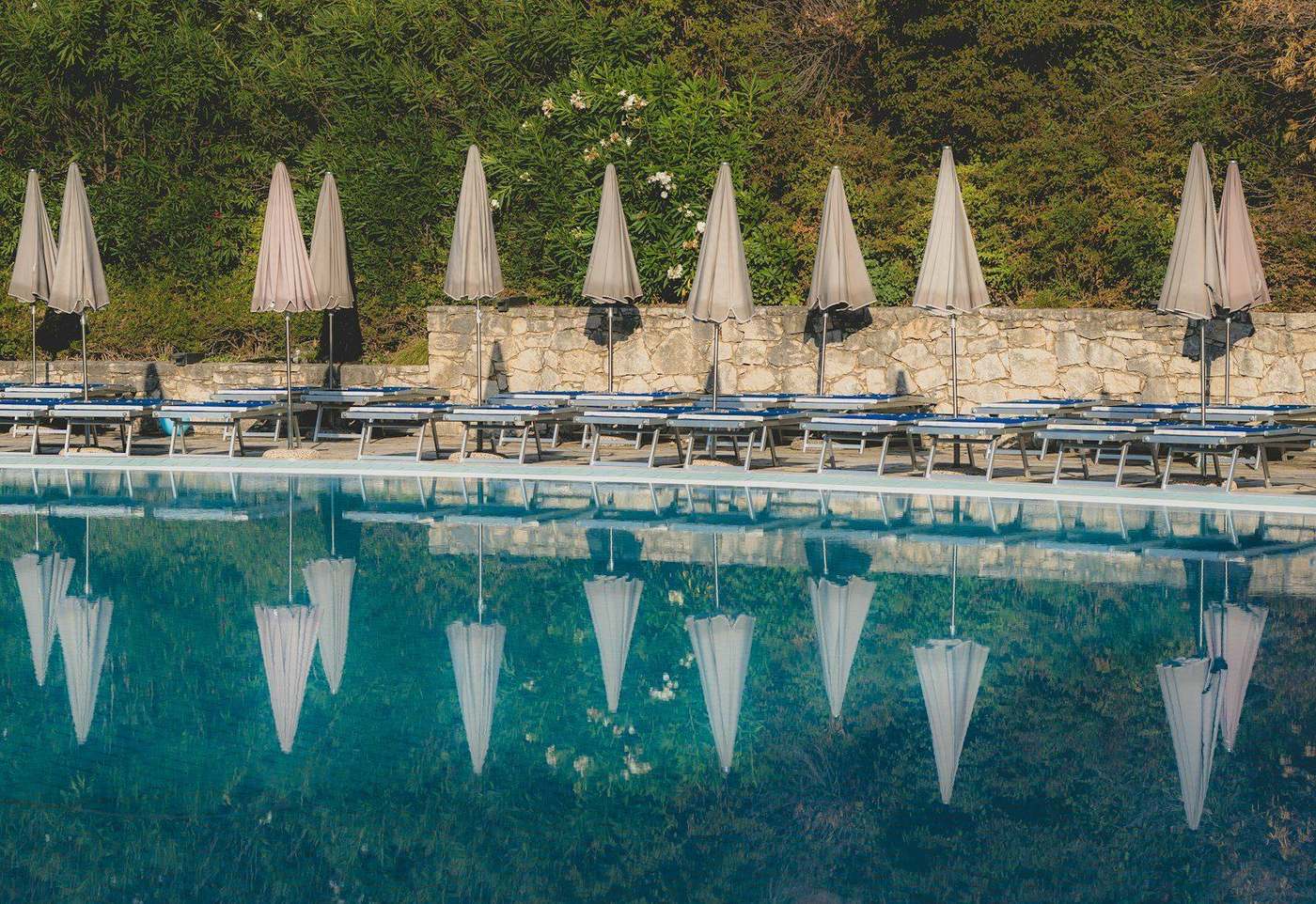 Piscina di uno stabilimento balneare con gli ombrelloni bianchi chiusi riflessi nell'acqua, i lettini allineati sul bordo e il muretto a secco con gli oleandri
