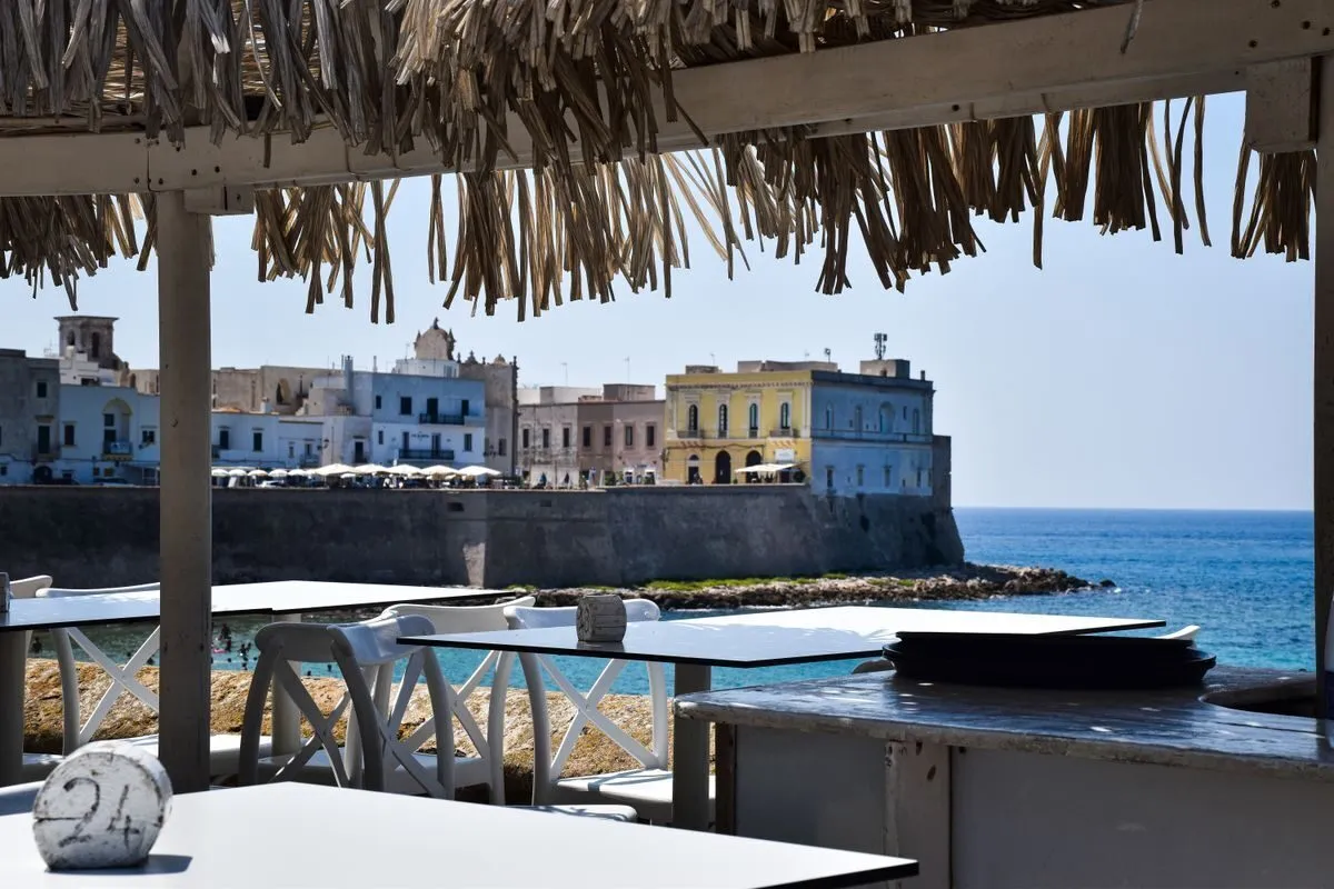 Tettoia di paglia di un lido salentino con i tavoli affacciati sulla città bianca e sul mare